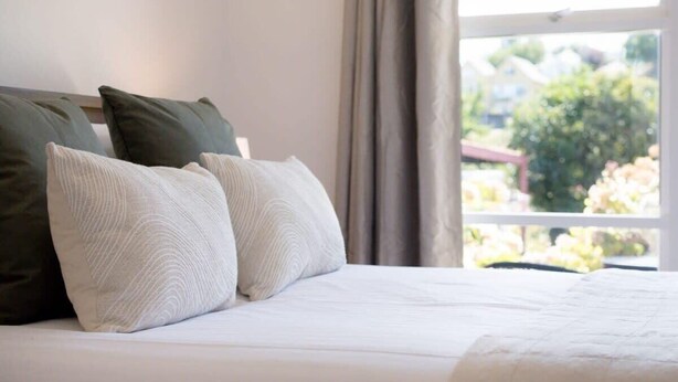 Una cama con ropa blanca y dos almohadas, una verde y otra blanca, en una habitación con ventana y cortinas.