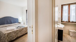 Un dormitorio con cama, mesita de noche, cortinas en la ventana y baño con lavabo y cesta.