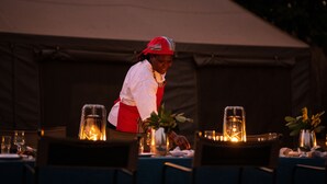 A person in a red and white uniform preparing a table with lit lanterns and a plant. - Nkula Walking Camp by RETURN Africa (Mutale)