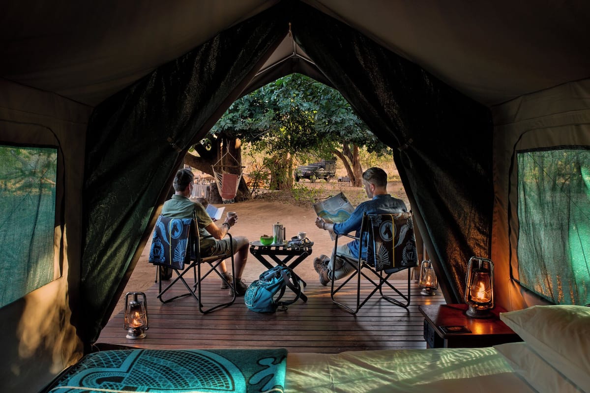 A tent with two people seated at a table, reading, with a view of a vehicle and trees outside.