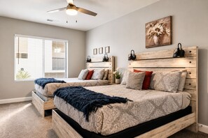 A bedroom with two beds, a ceiling fan, a window with blinds, a wall-mounted lamp, and a floral painting.