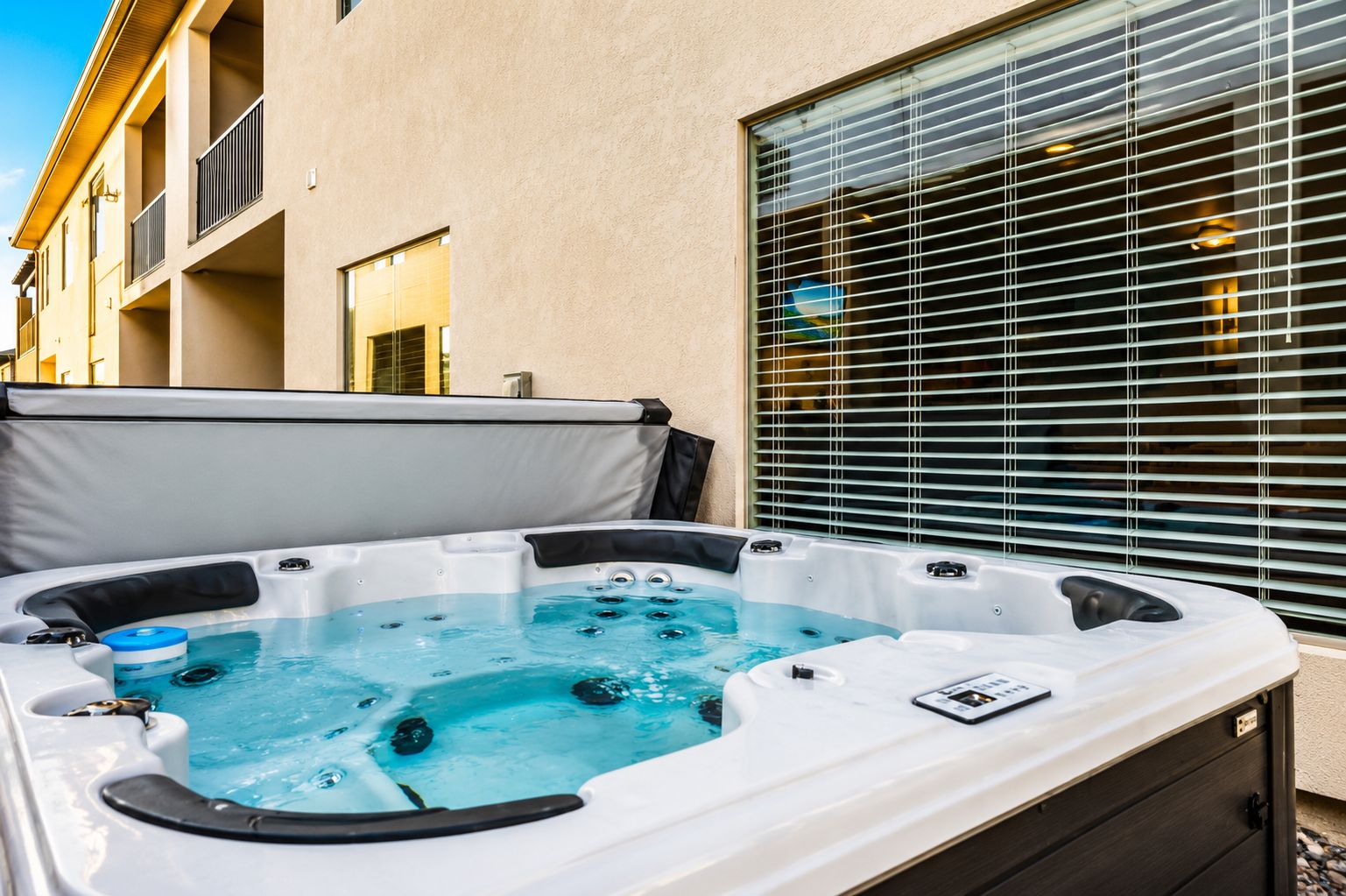 Un bain à remous doté d’un panneau de commande, installé devant un bâtiment avec une fenêtre et des stores.