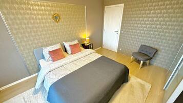 A bedroom with a large bed, a bedside table with a lamp, a chair, and a door.