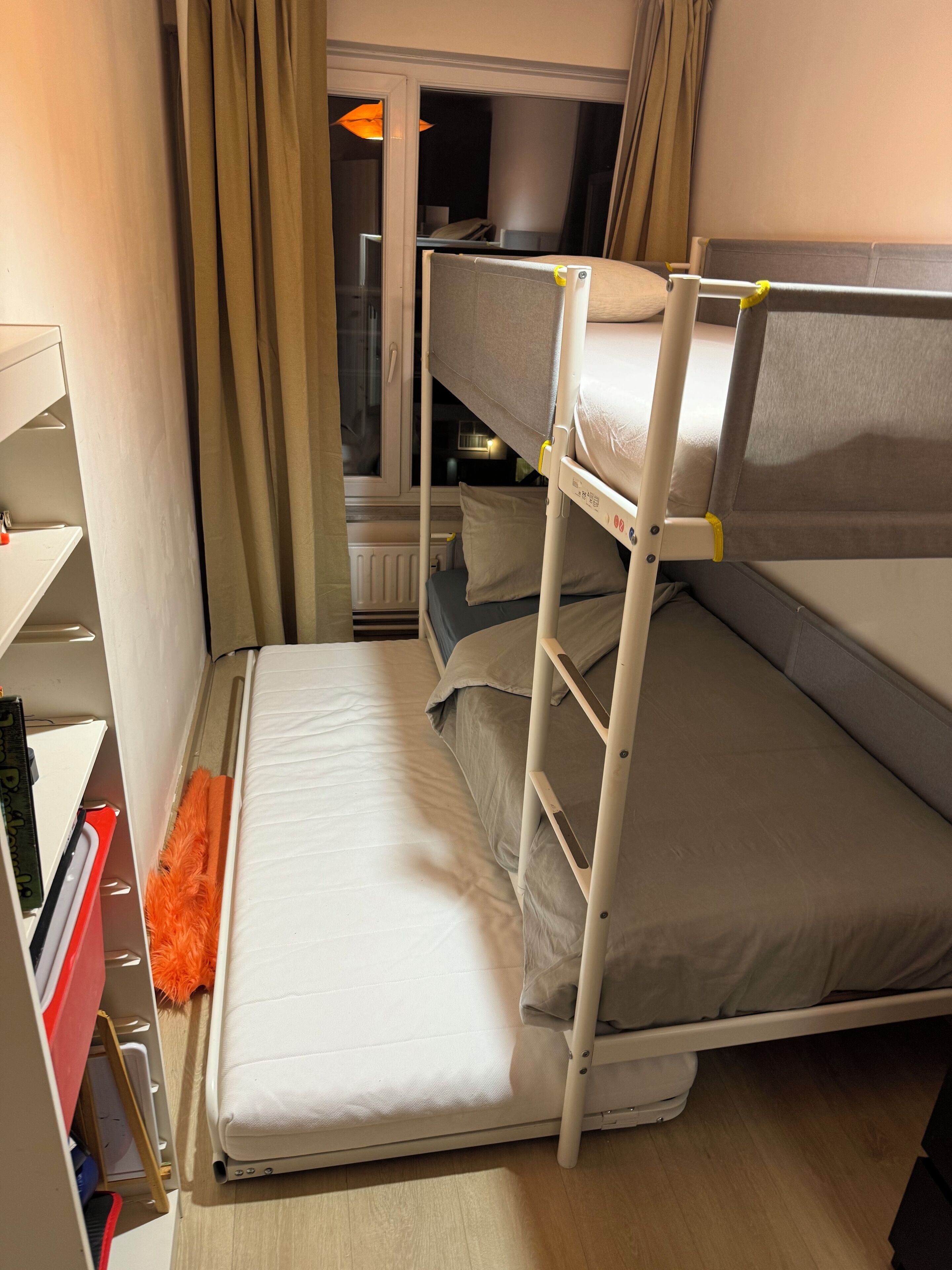 Pieni hotellihuone, jossa on kerrossängyt, pieni ikkuna, jossa on verhot, ja yöpöytä, jossa on lamppu.