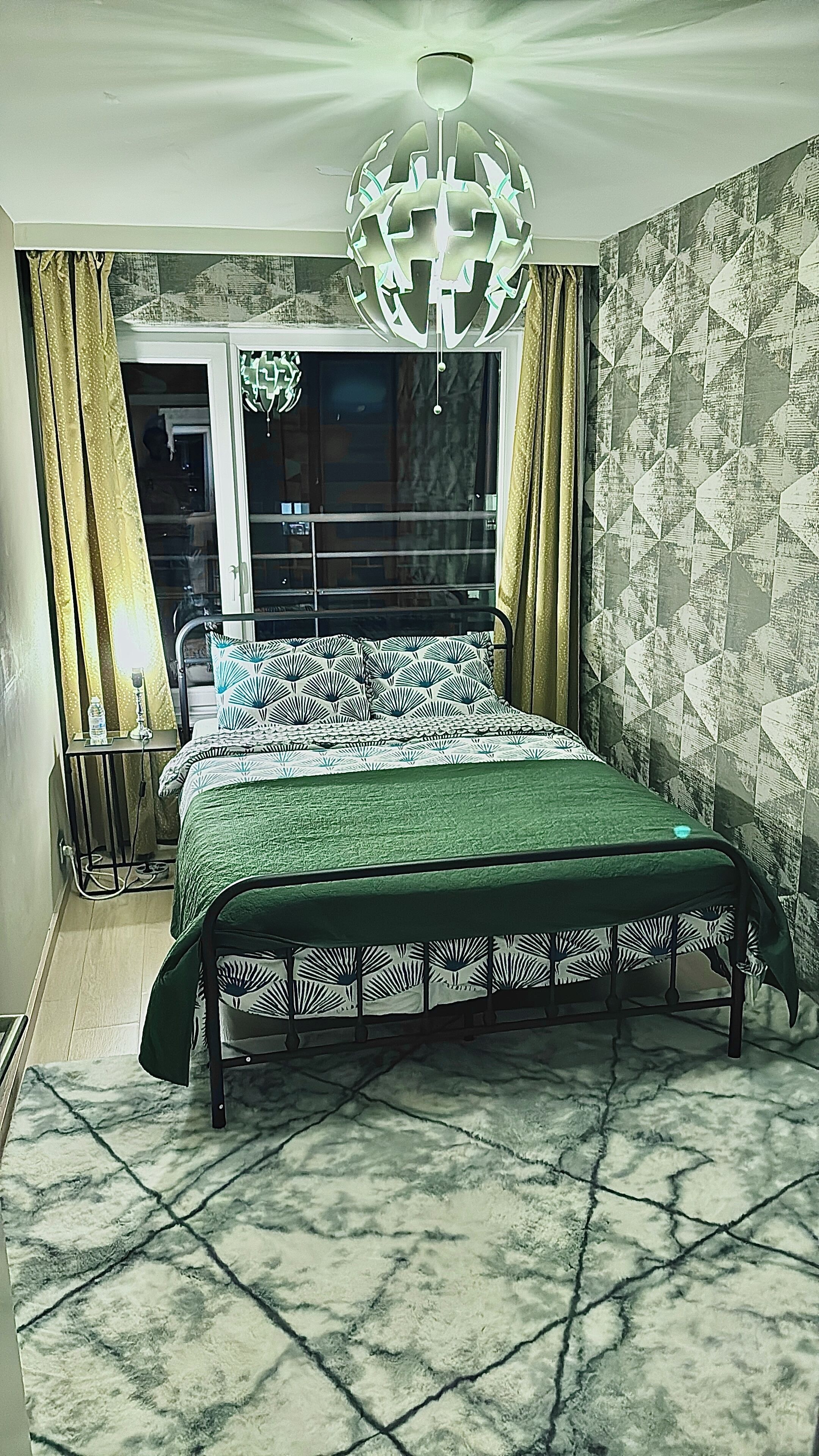 A bedroom with a bed, a chandelier, a window with curtains, and a patterned wall.
