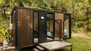 A small, modern cabin with a wooden exterior, large glass windows, and a wooden deck. - Natural Age by Tiny Away (Bahrs Scrub)