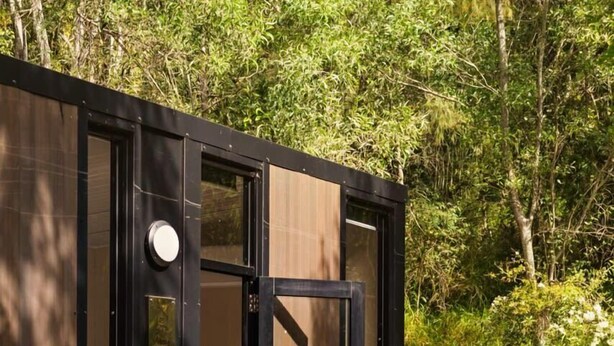 A small, modern cabin with a wooden exterior, a sliding door, and a wooden deck leading to a bed. - Natural Age by Tiny Away (Bahrs Scrub)