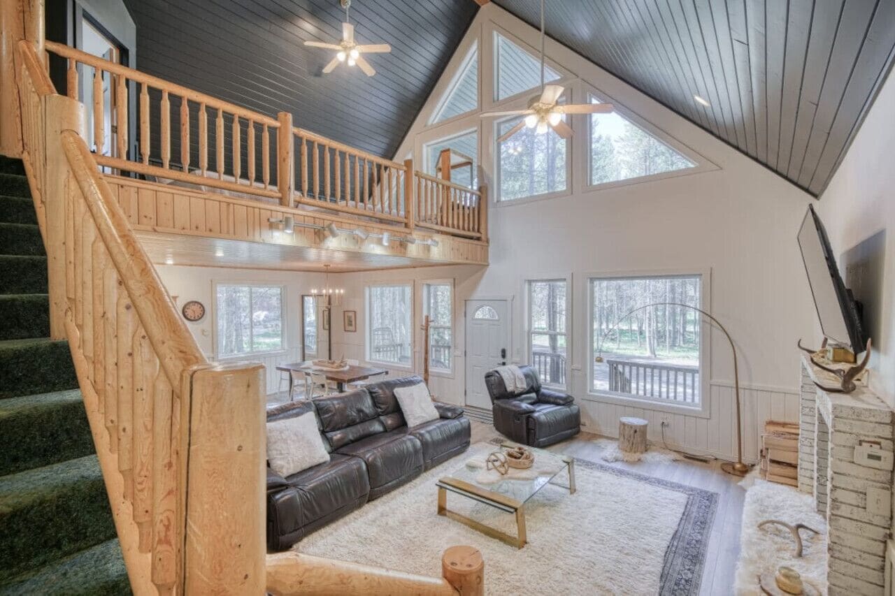A spacious living area with a high ceiling, a wooden staircase, a black leather sofa, a coffee table, and a television.