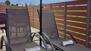 A patio area with two lounge chairs under a canopy, a metal fence, and a view of a cultivated field. - KaVros Cozy Apartment with mountain views (Paralia Kourna)