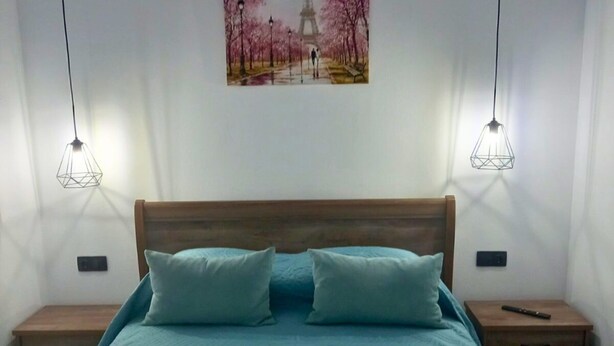 A bedroom with a large bed, two nightstands, a wooden headboard, and a painting of a park scene. - KaVros Cozy Apartment with mountain views (Paralia Kourna)