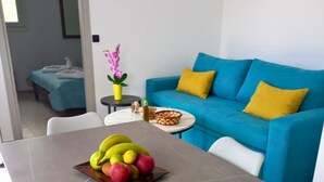 A modern living room with a blue sofa, a dining table with a fruit bowl, and a small side table with a vase and decorative items. - KaVros Cozy Apartment with mountain views (Paralia Kourna)