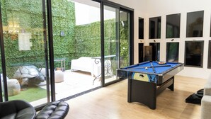 Una mesa de billar en una sala con ventanas amplias, un sofá y vistas a un jardín.