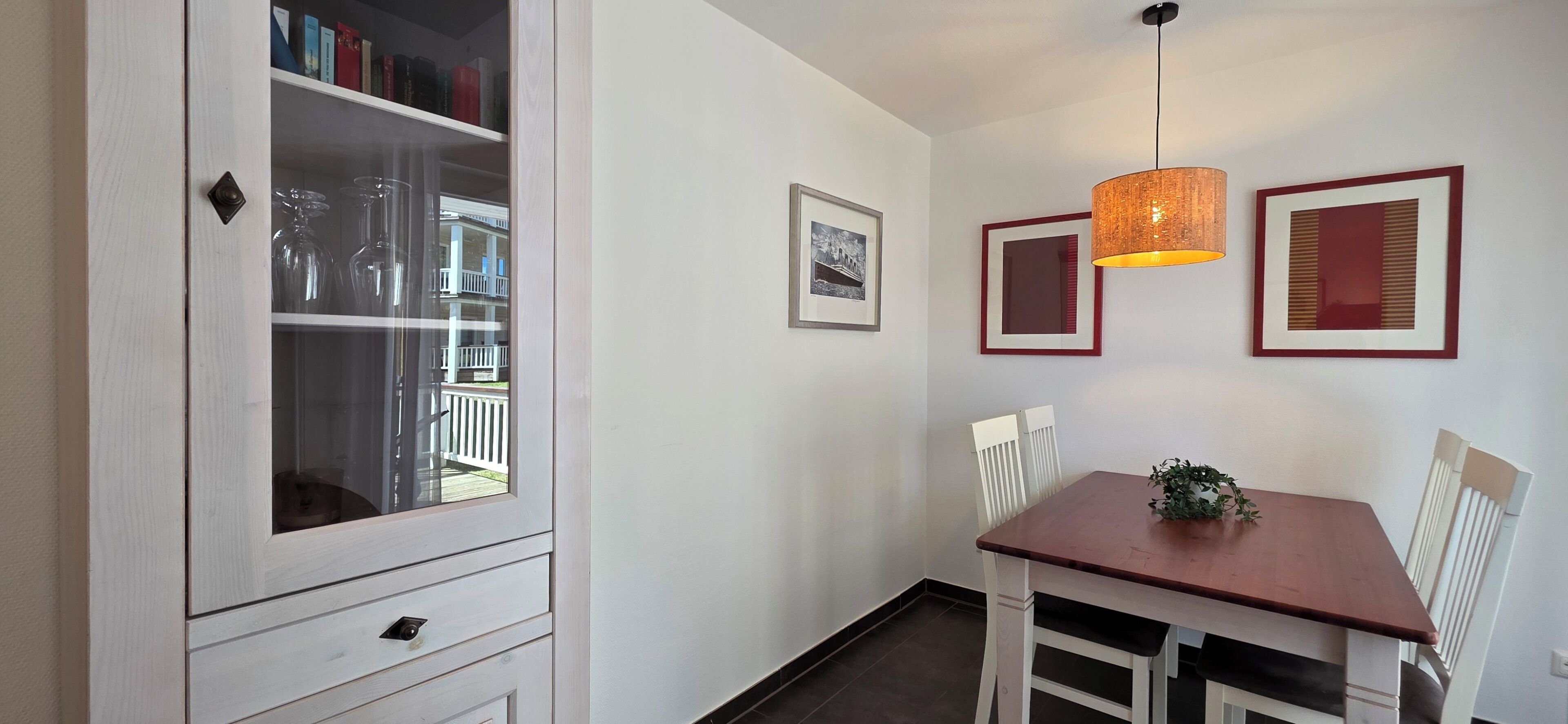 A dining area with a wooden table, white chairs, a hanging pendant light, and framed pictures on the wall.
