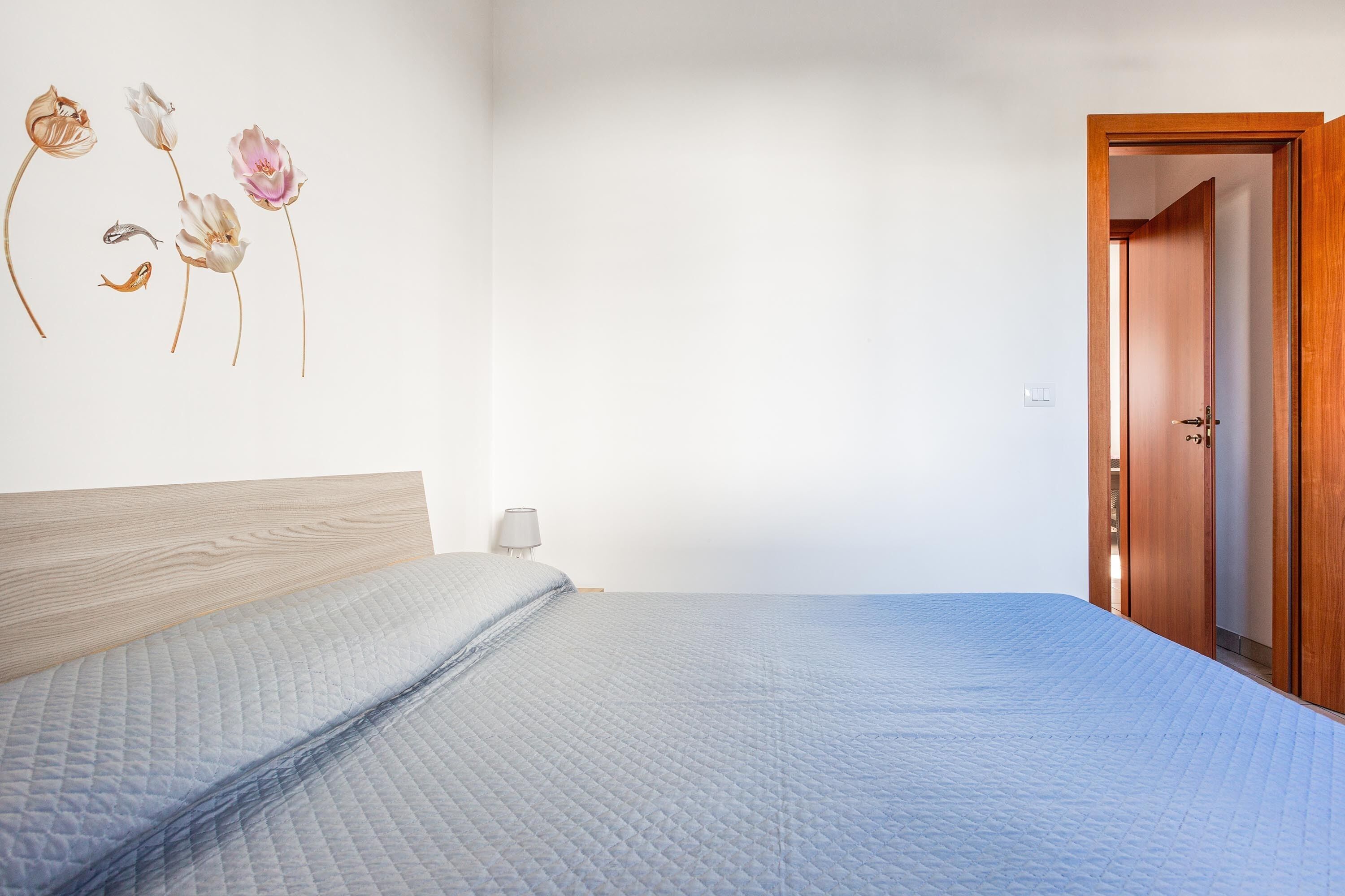 A bedroom with a wooden headboard, a quilted bedspread, a nightstand with a lamp, and a wall-mounted floral artwork.