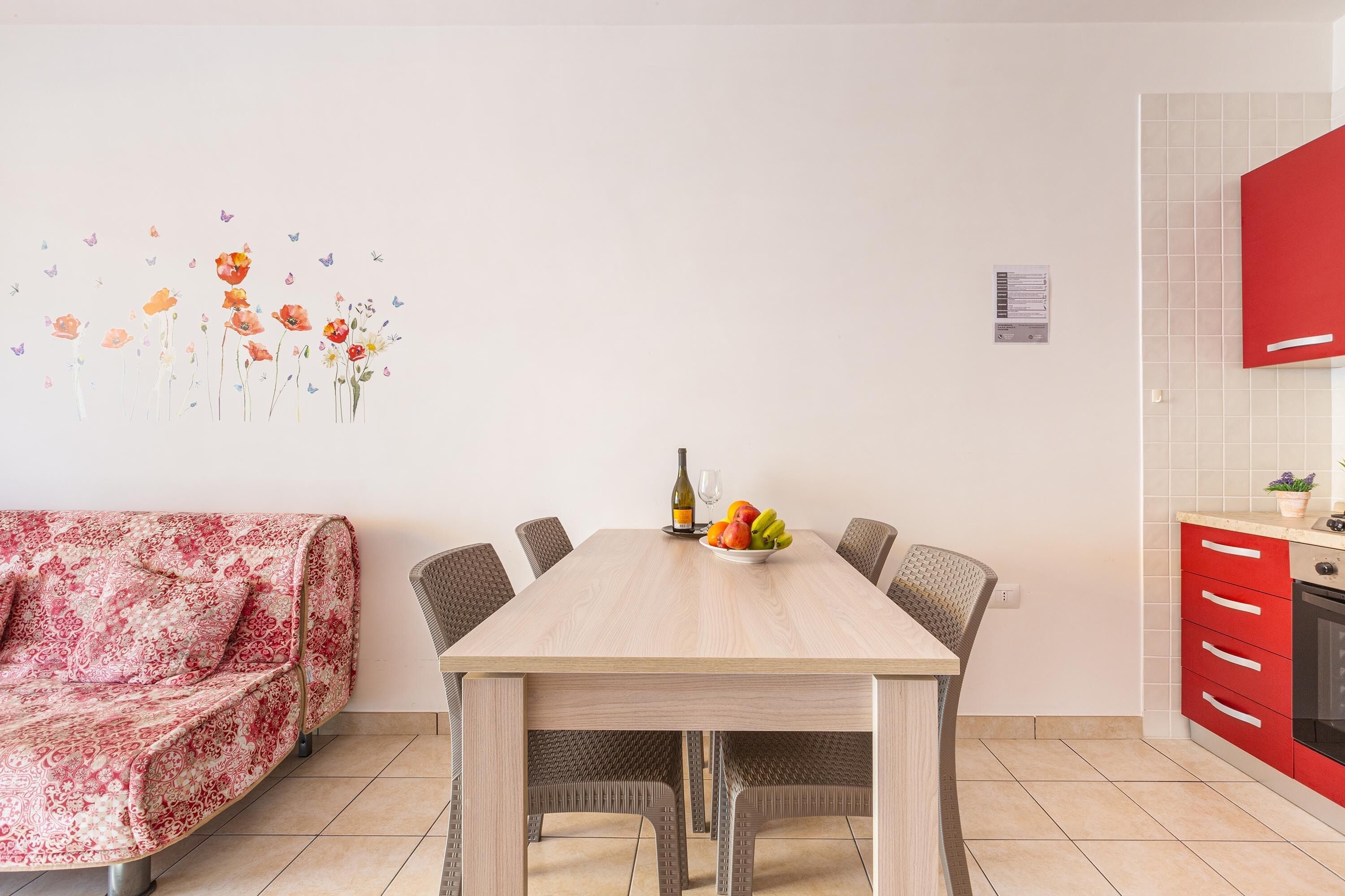 A dining area with a wooden table, six chairs, a red kitchen area, and a patterned sofa.