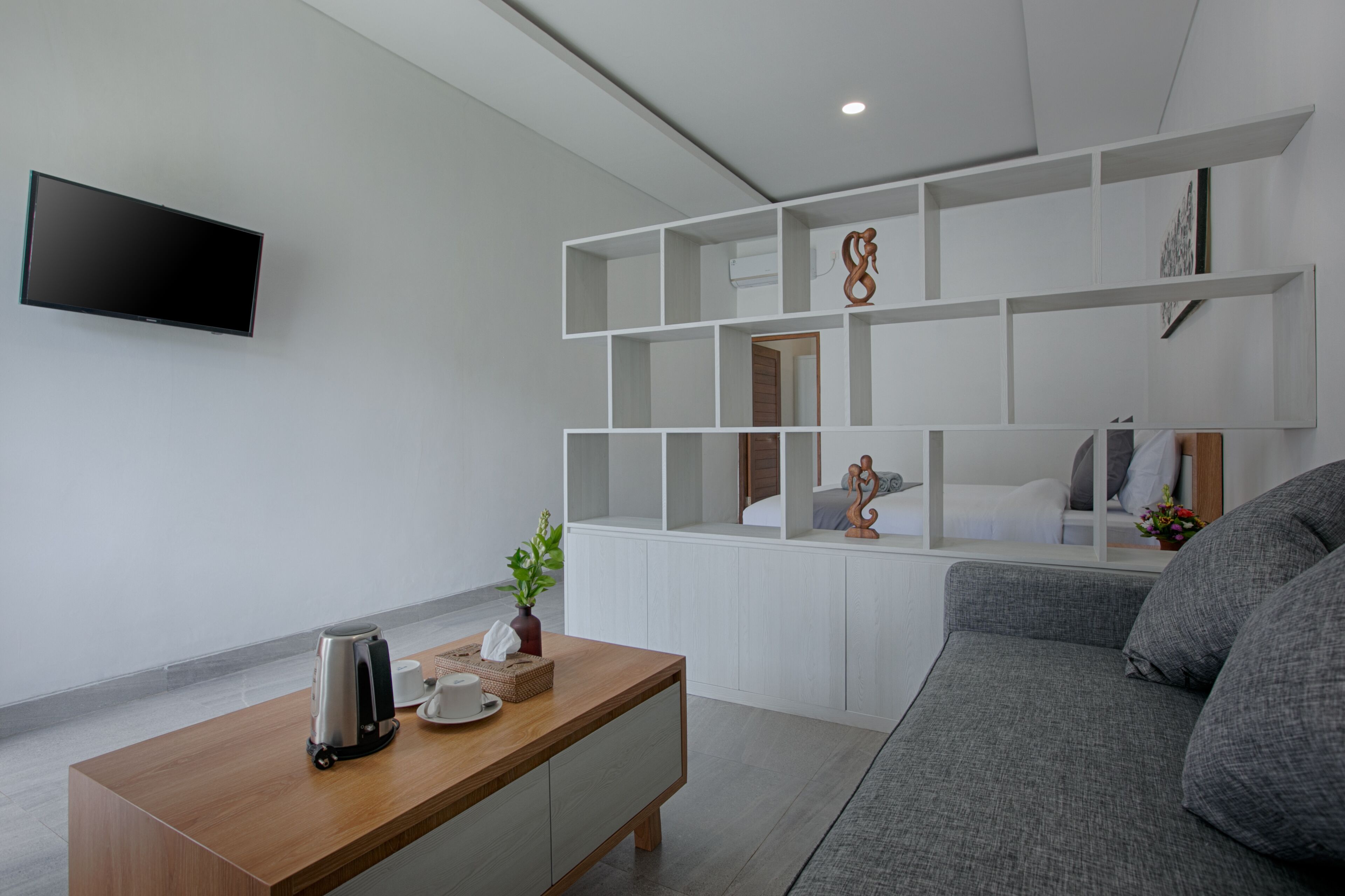 A modern living room with a flat-screen TV mounted on the wall, a wooden coffee table with a kettle and a vase, and a grey sofa.