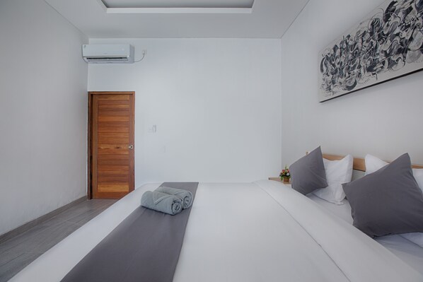 A modern hotel room with a wooden door, a bed with grey bedding and pillows, a small table with a vase, and a large abstract painting on the wall.