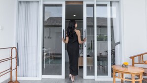 Une femme vêtue d’une robe noire se tient devant une porte-fenêtre coulissante. À sa droite, on aperçoit une chaise en bois et une petite table sur laquelle est posée une fleur jaune.