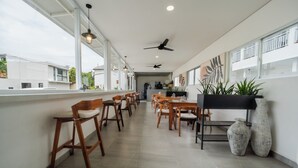 Un vaste espace dédié à la salle à manger, avec des chaises et des tables en bois, de grandes fenêtres, des ventilateurs de plafond et des plantes en pot.