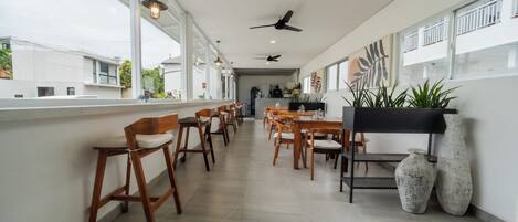 A spacious dining area with wooden chairs and tables, large windows, ceiling fans, and potted plants.