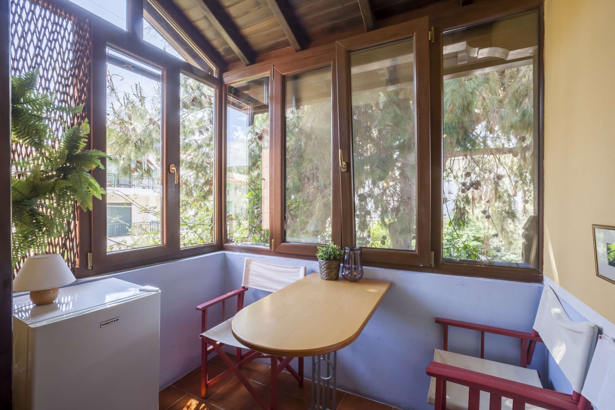 Habitación con techo de madera, una mesa con un banco, un refrigerador pequeño y ventanas amplias con vistas a árboles.