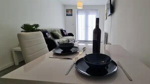 Un coin repas avec une table dressée pour deux personnes, un canapé, une plante en pot et une bouteille de vin.