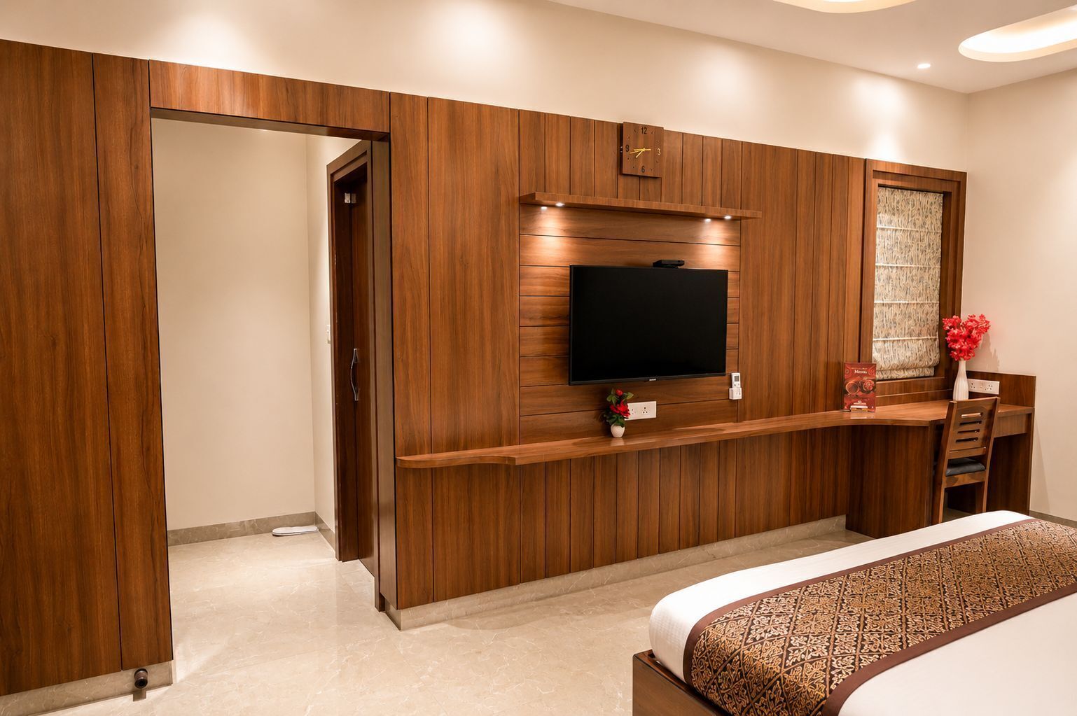 A bedroom with a wooden wardrobe, a flat-screen TV mounted on the wall, a bedside table with a vase of flowers, and a bed with patterned bedding.