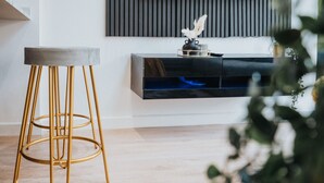 A modern living room with a wooden floor, a grey cushioned stool, a black console table with a decorative item, and a black shelving unit.