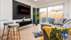 A modern living room with a flat-screen TV mounted on a black panel, a grey sofa with patterned cushions, a yellow throw blanket, and a potted plant.