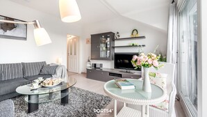 Uma sala de estar moderna com sofá, mesa de centro e uma área de jantar com mesa de vidro. Há uma cozinha com televisão e armários.
