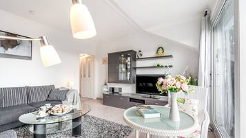 A modern living room with a sofa, coffee table, and a dining area with a glass table. There is a kitchen with a TV and cabinets.