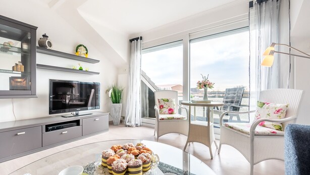 Uma sala de estar moderna com uma TV de tela plana, uma mesa de jantar com arranjo floral, cadeiras de vime e uma janela grande com vista.