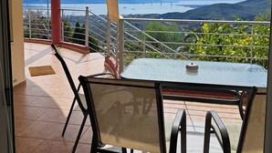 Een balkon met een glazen tafel en stoelen, met uitzicht op een schilderachtig landschap met bergen en een brug.