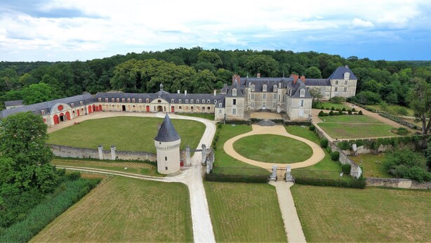 A large, historic building with a central courtyard, surrounded by manicured gardens and a forested hill.