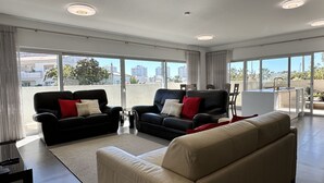 Amplia sala de estar con dos sofás de cuero negro, un sofá beige y una mesa de comedor de vidrio. Amplias ventanas ofrecen vistas a la ciudad.