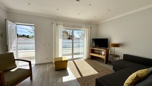 Un salon moderne avec un canapé, un téléviseur posé sur un meuble en bois et une grande porte-fenêtre coulissante donnant sur un balcon extérieur.