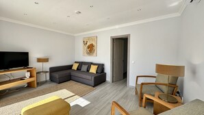 Un salon moderne comprenant un canapé d’angle, un téléviseur à écran plat, un meuble de rangement en bois et un coin repas avec une table et des chaises.