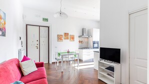 En moderne stue med en rød sofa, et spiseområde med et grønt bord og et køkken med hvide skabe samt et fjernsyn.