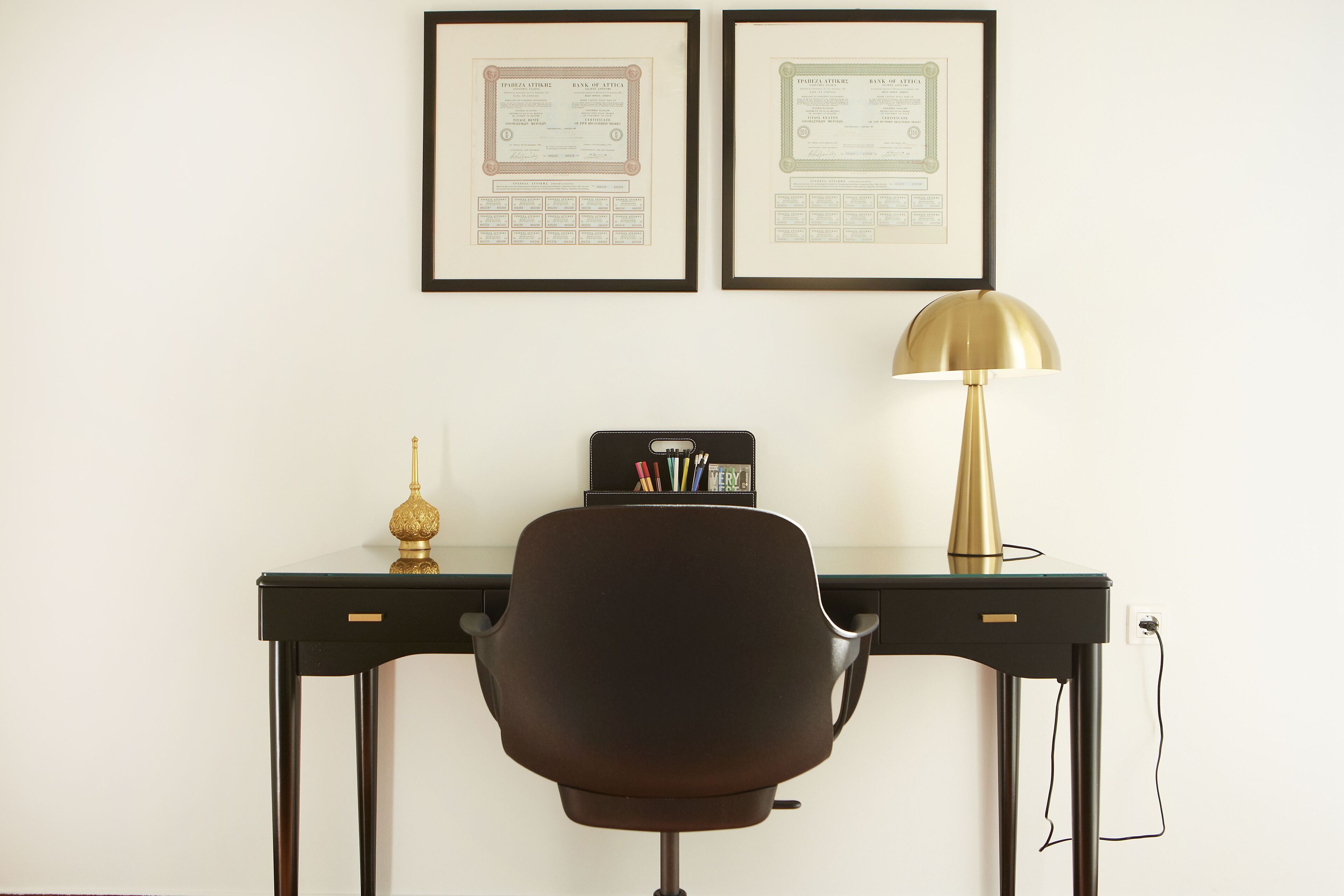 A desk with two framed certificates, a black office chair, a gold desk lamp, and a black desk with a pen holder.