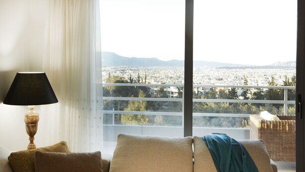 A room with a sofa, a lamp, and a view of a cityscape through a large window. - Luxury Apartment with Exceptional View (Papagou-Holargos)