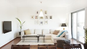 A modern living room with a sofa, coffee table, and chairs. There is a flat-screen TV mounted on the wall, a lamp, and a large window providing natural light. - Luxury Apartment with Exceptional View (Papagou-Holargos)
