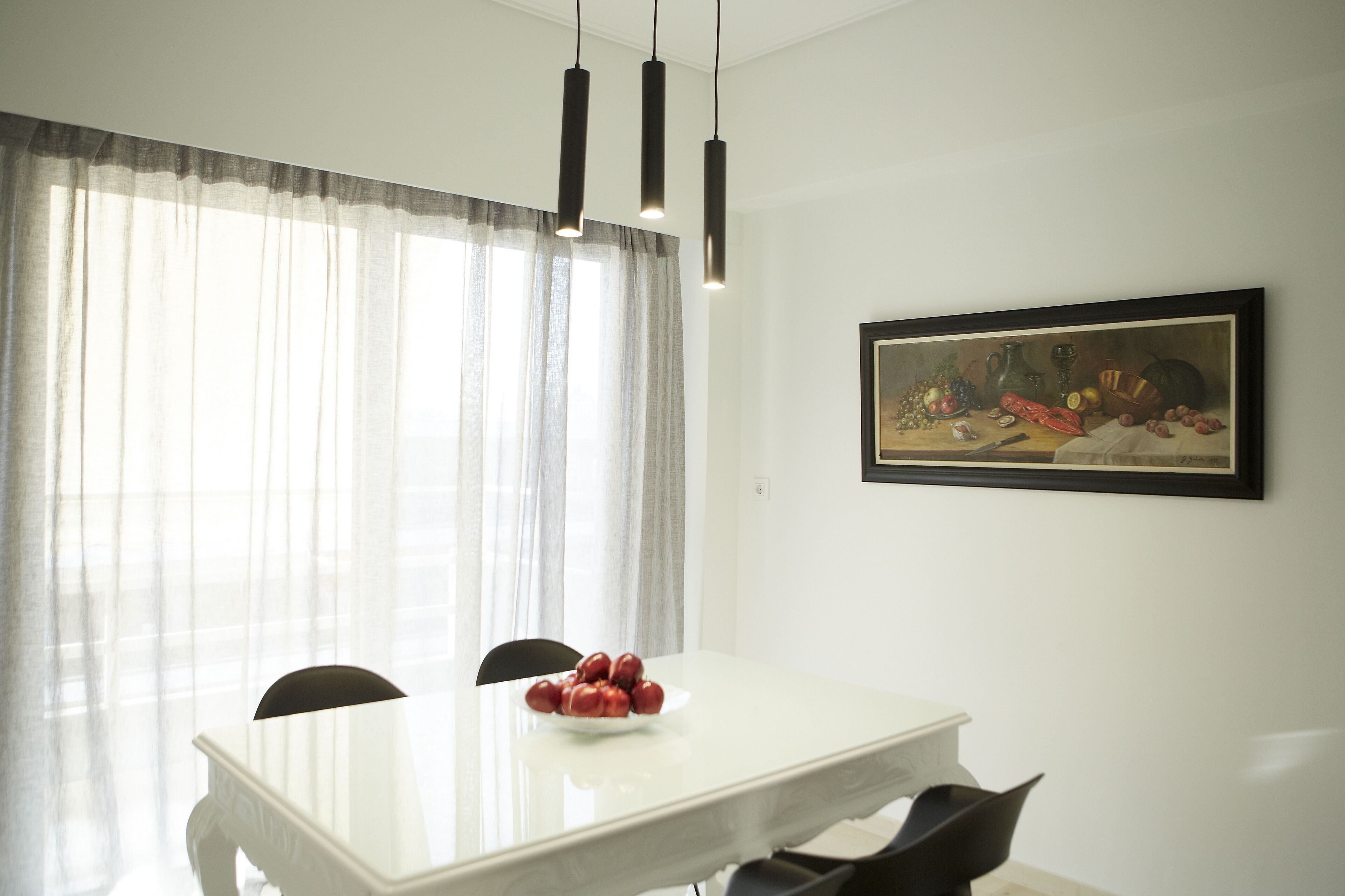 Un coin repas avec une table blanche, des chaises noires, un saladier de fruits, un tableau au mur et une fenêtre ornée de rideaux légers.