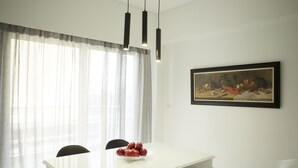 A dining area with a white table, black chairs, a bowl of fruit, a painting on the wall, and a window with sheer curtains. - Luxury Apartment with Exceptional View (Papagou-Holargos)