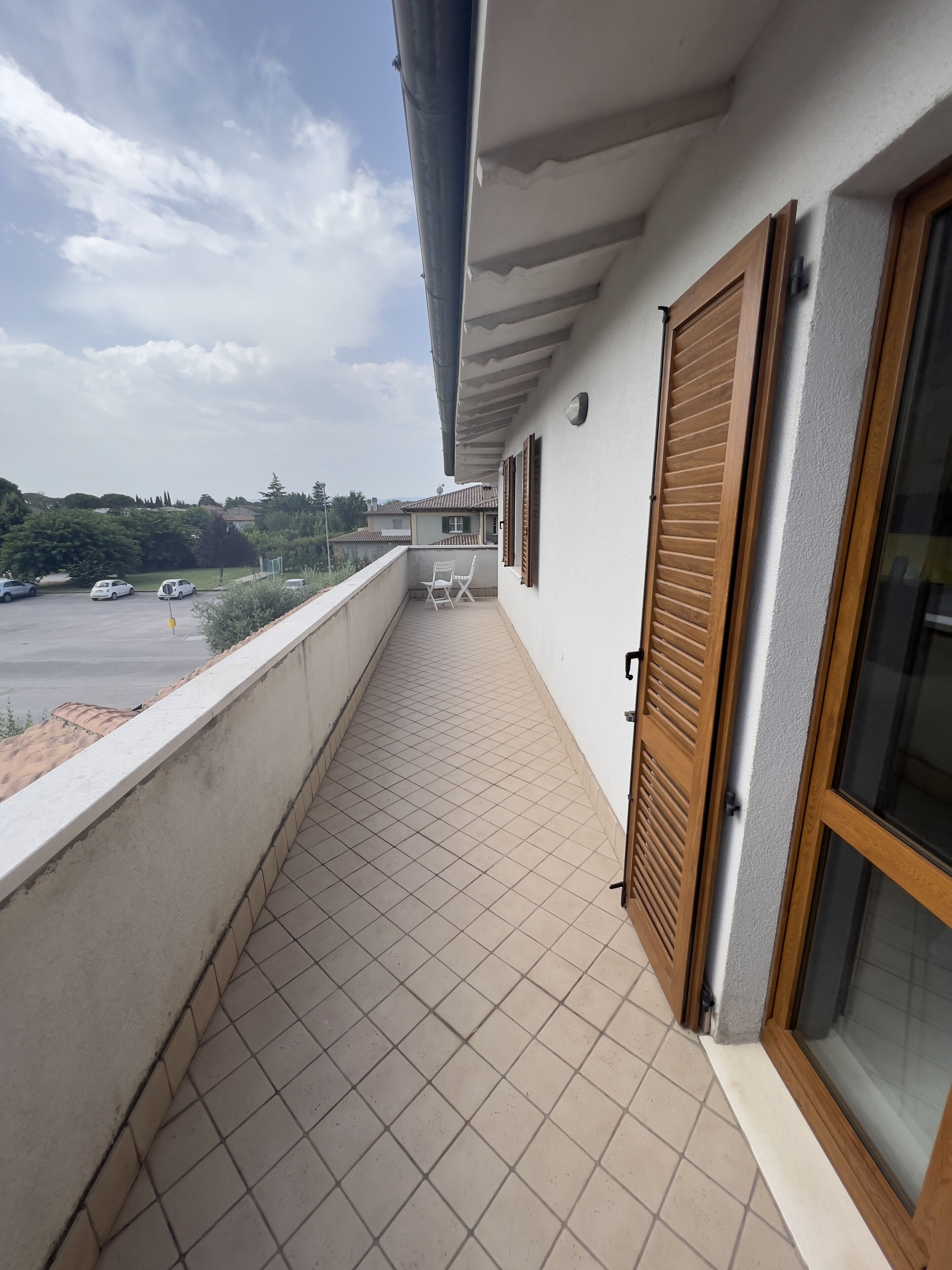A balcony with tiled flooring, a chair, and a view of a parking lot and distant buildings.