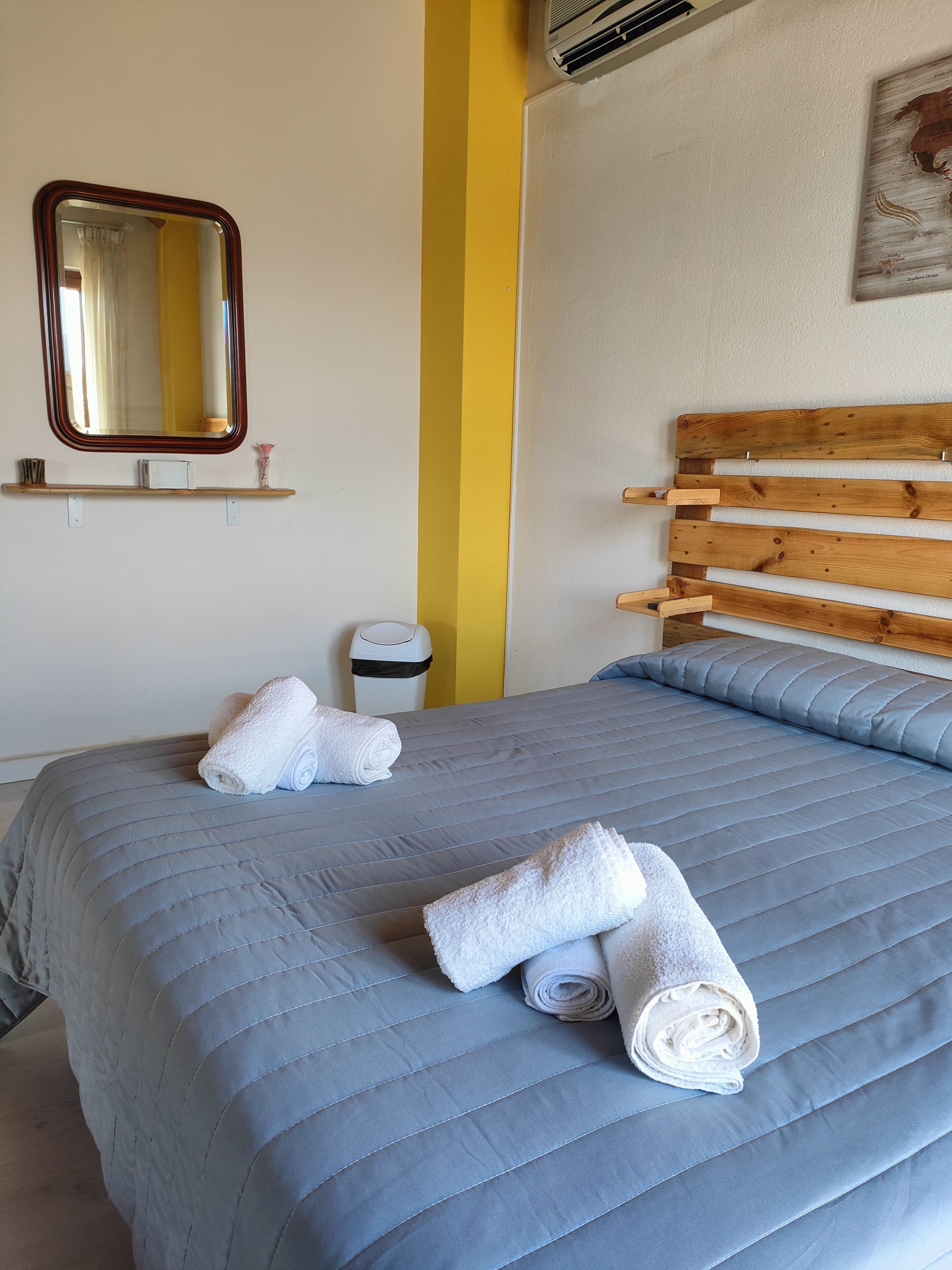 A neatly made bed with a blue quilt and two rolled white towels, a wooden headboard, a small shelf with items, a mirror, and a wall-mounted air vent.