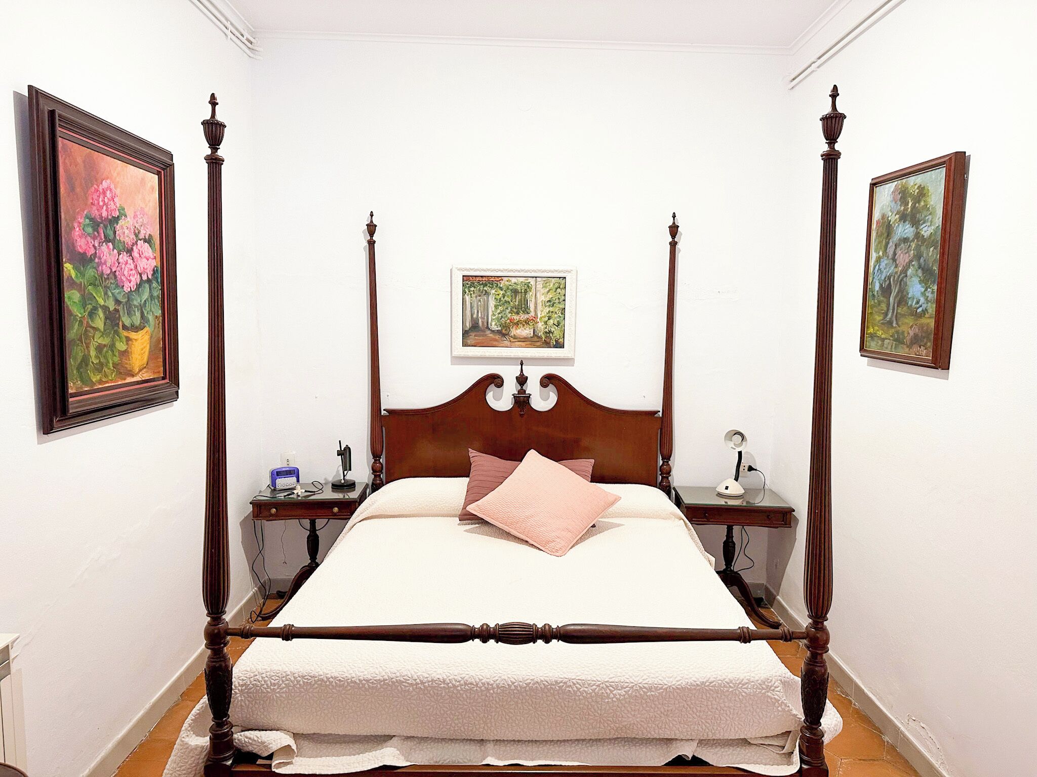 Een hemelbed met houten hoofdeinde en voeteinde, omringd door twee nachtkastjes met lampen, in een kamer met witte muren en bloemschilderijen in lijsten.