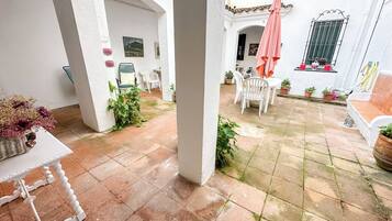 Een binnenplaats met witte muren, betegelde vloeren en potplanten. Er staan witte stoelen en een tafel, en een roze parasol.