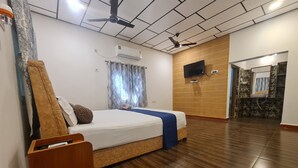 A hotel room with a wooden floor, a bed with a blue blanket, a ceiling fan, an air conditioning unit, and a television on the wall. - Dev Farm Sasan Gir (Talala)