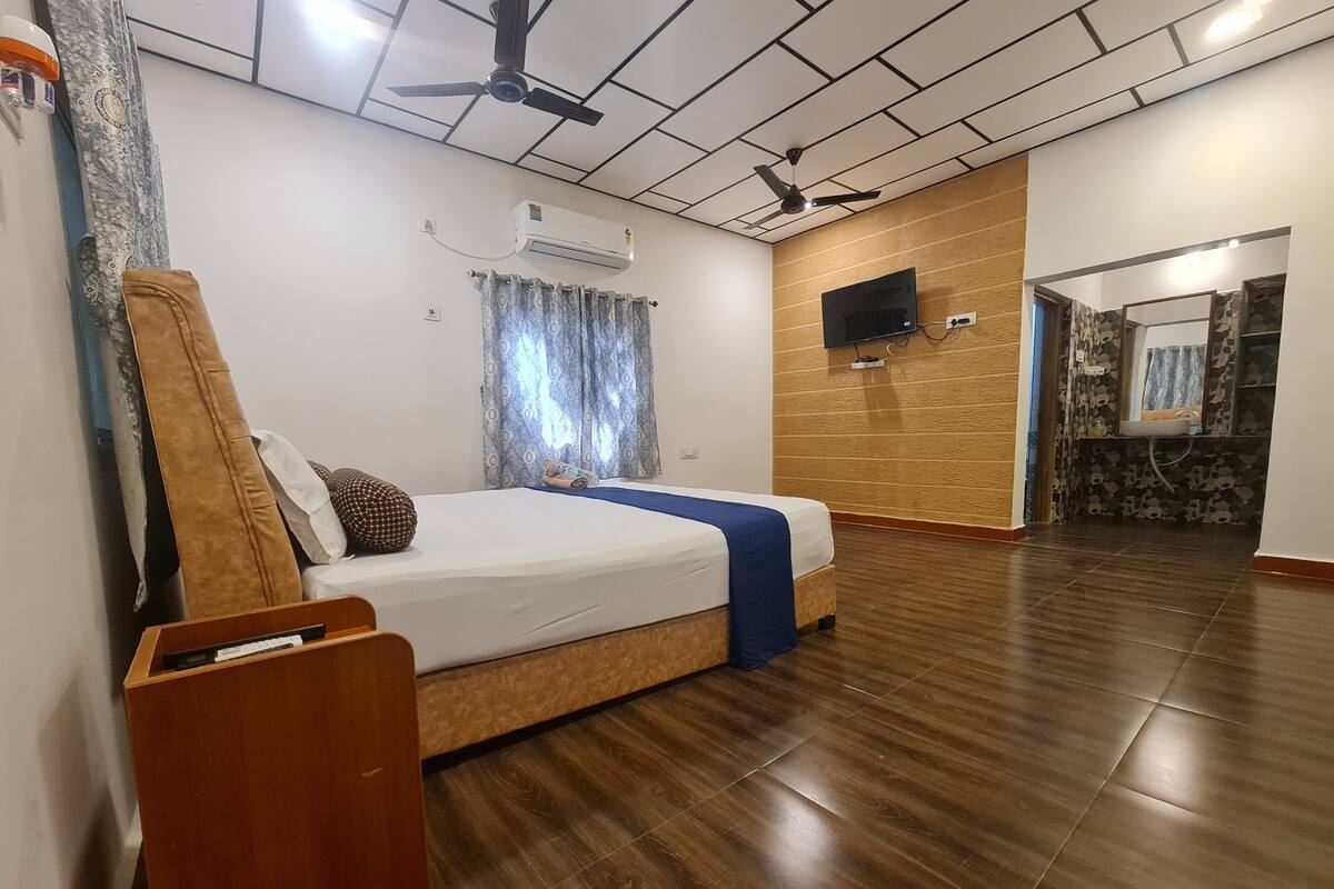 Habitación de hotel con piso de madera, cama con manta azul, ventilador de techo, aire acondicionado y televisor en la pared.
