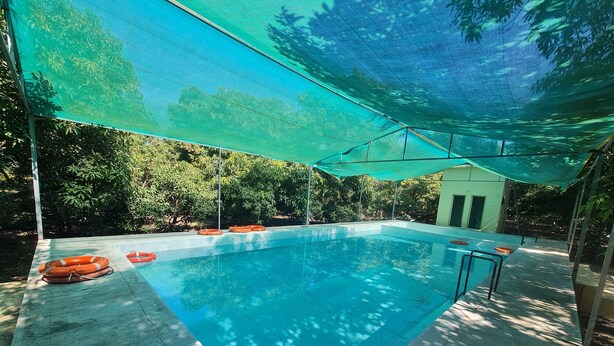 A swimming pool with a shaded area, surrounded by greenery and a building in the background. - Dev Farm Sasan Gir (Talala)